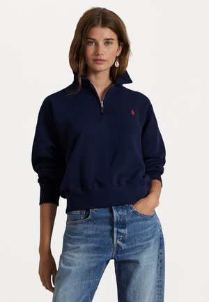 Marineblauwe zip-up sweatshirt met een hoge kraag, geribbelde manchetten en een klein rood logo op de borst, gecombineerd met lichtblauwe distressed jeans.