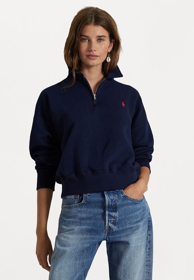Marineblå zip-up sweatshirts med høj krave, ribbede manchetter og et lille rødt logo på brystet, kombineret med lyseblå slidte jeans.
