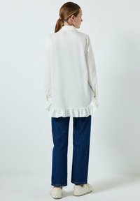 Camisa blanca de manga larga con volante en el bajo, combinada con pantalones vaqueros de pierna ancha en azul oscuro y zapatillas blancas. Textura suave, diseño minimalista.