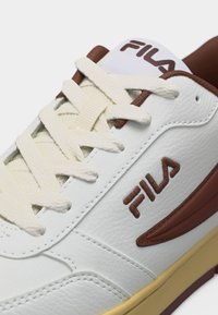 Λευκό αθλητικό παπούτσι FILA με εκρού κορδόνια, καφέ ραφή λογότυπου, καφέ εσωτερική επένδυση και μπεζ σόλα, εμφανιζόμενο σε κοντινή λήψη λεπτομέρειας.