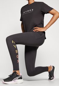 Svart atletisk t-shirt med "HYROX" logo, ihopparad med svarta leggings med färgglad lettering. Svarta och rosa löparskor fulländar looken.