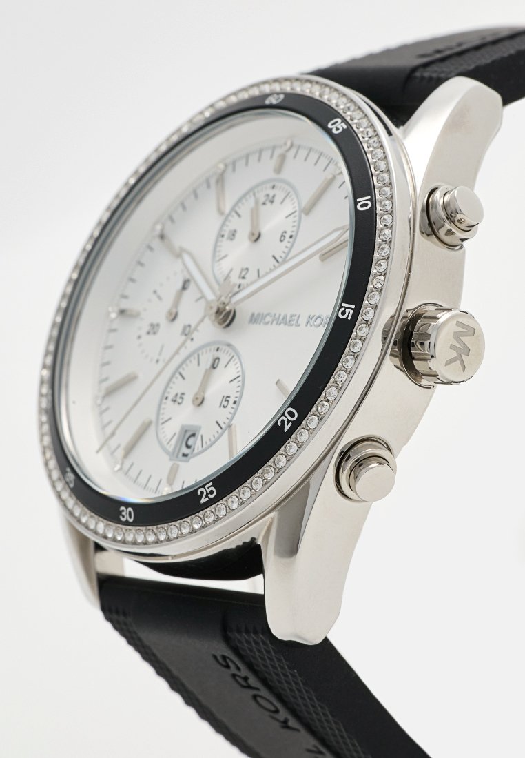 Silberfarbene Uhr mit weißem Zifferblatt, Chronographen-Subdials, schwarzem Lünette und Gummiarmband. Besondere Merkmale sind Kristallakzente auf der Lünette und dem Logo.