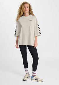 Overmaat lichtbeige t-shirt met zwart logo en diagonale strepen op de mouwen, gecombineerd met zwarte leggings en gestreepte sokken, afgewerkt met sneakers.