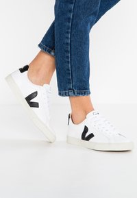 White sneakers with black accents, made of smooth leather featuring a logo detail. Rubber sole and laces, paired with dark denim jeans.