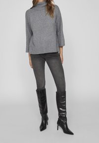 Grau gestrickter Pullover mit hohem Kragen, dreiviertel langen Ärmeln, getragen mit dunkelgrauen Skinny-Jeans und kniehohen schwarzen Lederstiefeln.