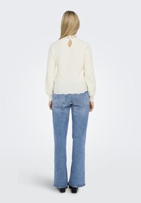 White knitted sweater with a scalloped hem, featuring a keyhole cutout at the back. Paired with light blue flared jeans.