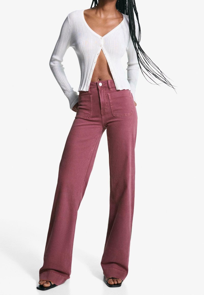 Personne portant un cardigan court côtelé blanc et un pantalon large taille haute mauve avec poches plaquées à l'avant ainsi que des sandales noires à bout ouvert.