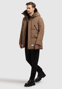 Hnedá prešívaná bunda s kapucňou, viacerými zipsovými vreckami a prešívanou textúrou. Model nosí čierne nohavice a robustné čierne topánky.