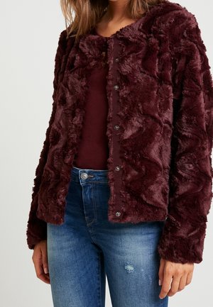 Woman wearing a dark burgundy faux fur button-up jacket over a matching top with mid-blue distressed jeans against a plain background.