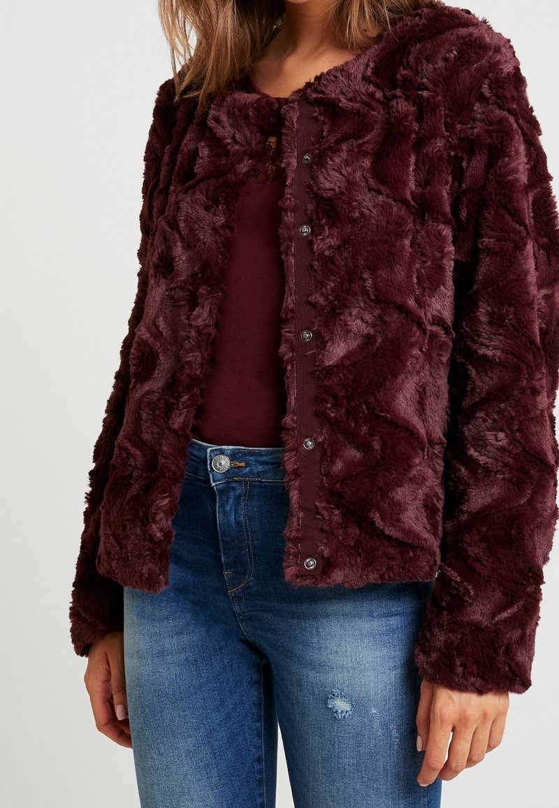 Femme portant une veste boutonnée en fausse fourrure bordeaux foncé sur un haut assorti avec un jean bleu moyen déchiré sur un fond uni.