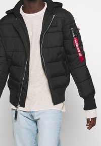 Sort puffet jakke med lynlås, ribbede manchetter og hætte. Har et rødt stofmærke på venstre ærme og et quiltet design.