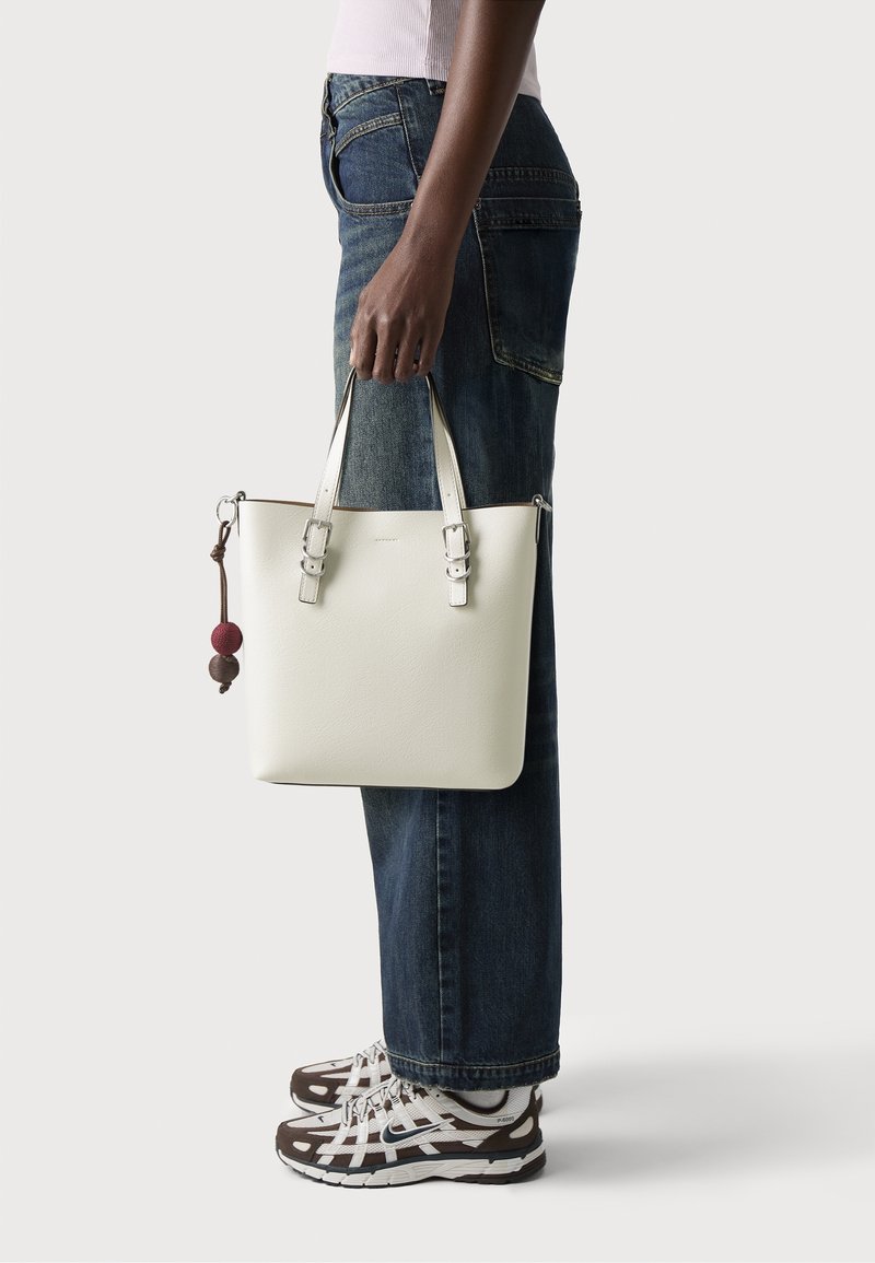 Personne en jean bleu et baskets tenant un sac à main en cuir blanc avec double poignée et deux perles décoratives sur le côté.