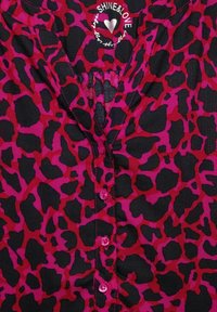 Blusa nera trasparente con stampa leopardata rosa e rossa. Presenta un collo a V, bottoni lungo il fronte e un logo a forma di cuore sul colletto.