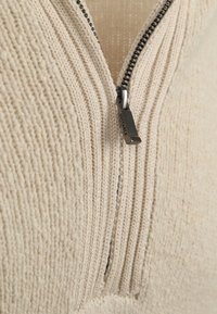 Close-up van beige geribbelde gebreide stof met een gedeeltelijk open ritssluiting waarvan het zwarte trekstuk is gelabeld met "SUITABLE".