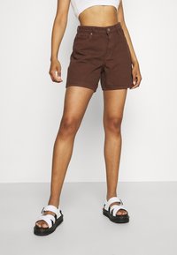 Pantalones cortos de algodón marrón con frente plano, dos bolsillos y cierre de botón, combinados con sandalias blancas de tiras sobre un fondo blanco.