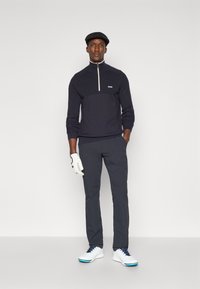 Pullover azul-marinho com fecho de zíper branco e logótipo, combinado com calças antracite e sapatos de golfe brancos com detalhes azuis. Luva de golfe branca na mão esquerda.