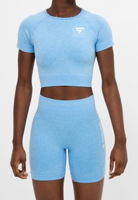Žena v světle modrém těsném krátkém topu s krátkými rukávy a vysokými cyklistickými šortkami s bílými logy na hrudi a stehně.