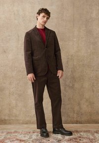 Brauner Cordanzug mit einem Blazer mit Spitzlapel und passenden Hosen, kombiniert mit einem roten Pullover und schwarzen Schnürschuhen. Strukturierten Stoff.