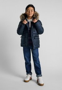 Marinblå puffjacka med fuskpälsfodrad huva, blixtlås fram, sidofickor; bärs över en randig polotröja och blå jeans, ihop med vita sneakers.
