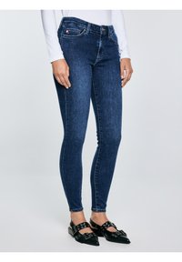 Naine, kes kannab tumelisi siniseid kitsaid teksapükse ja musti teravate ninadega kingi, millel on pandla ja silmkoe detailid, seisab helehallil taustal.