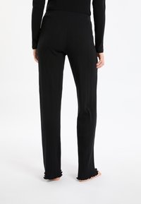 Persoon in zwarte ribgebreide broek met een lichte flare aan de zoom, staat blootsvoets, van achteren gezien op een witte achtergrond.