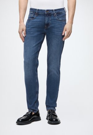 Homme portant un jean bleu slim et des mocassins en cuir noirs, debout devant un fond clair uni.