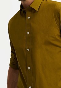 Oliivinvihreä paita, joka on valmistettu sileästä kankaasta. Siinä on tavallinen kaulus, pitkät hihat ja etupuolella nappilista pyöreillä napeilla.