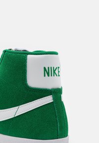 Gröna mocka högtoppsneakers med vita läderdetaljer och Nike-logotyp. Har en kontrasterande vit swoosh och strukturerad yttersula.