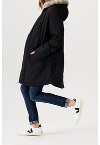 Černá parka bunda s kapucí ozdobenou umělým kožešinovým lemem. Nošeno s tmavě modrými džínami a bílými teniskami s černými detaily.