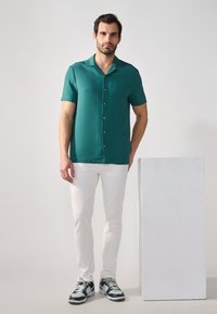 Tea zöld, rövid ujjú ing textúrázott mintával és gombos elülső résszel, fehér, slim-fit nadrággal és sötét sportcipővel párosítva. Egyszínű háttér.