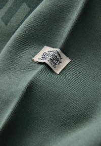 Zaļš kokvilnas audums ar tekstūru, kas parāda austu rakstu; ietver kvadrātveida austu etiķeti ar melnu tekstu, kas lasāms "Superdry."