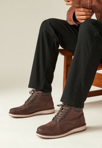 Hnedé kožené členkové topánky s čiernymi očkami na šnurovanie, šnúrkami a štruktúrovaným povrchom. Majú svetlú gumovú podrážku s prúžkovaným dizajnom.