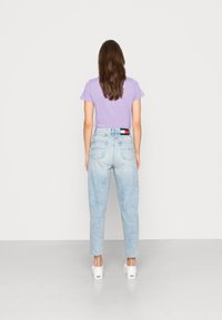 Kvinna med långt brunt hår som bär en lila t-shirt, ljusblå högmidjade jeans och vita sneakers, stående vänd bort.