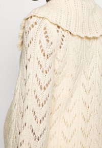 Stickad cardigan i krämfärg med en stor krage, som har ett chevronmönster och öppen vävstruktur. Mjuk, lätt material.