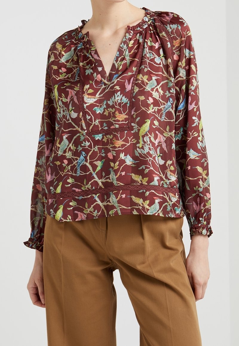 Blouse florale rouge avec un décolleté en V, des manches longues bouffantes et des détails plissés. Présente des motifs d'oiseaux colorés sur un tissu texturé.