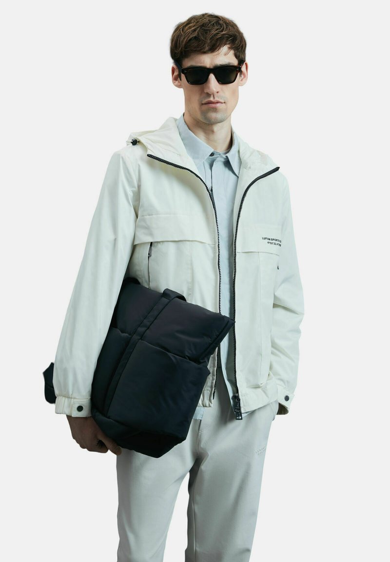 Veste imperméable blanche avec capuche, zips à l'avant et poches ; sac noir rembourré tenu d'une main ; chemise à col bleu clair ; pantalon gris.