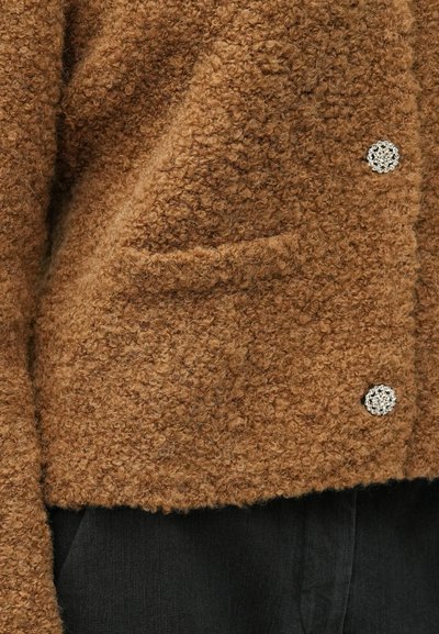 Brązowa teksturowana kurtka z puszystego materiału, z dwoma bocznymi kieszeniami i srebrnym ozdobnym guzikiem z przodu.