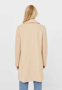 Abrigo de mezcla de lana beige con cuello de muesca, corte recto y mangas largas, que presenta un acabado texturizado y una abertura en la parte trasera para facilitar el movimiento.