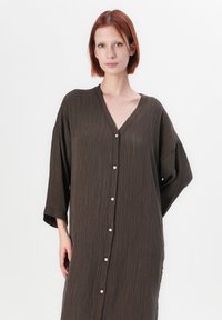 Femme aux cheveux roux courts portant une robe boutonnée ample et texturée de couleur marron foncé, sur fond blanc.