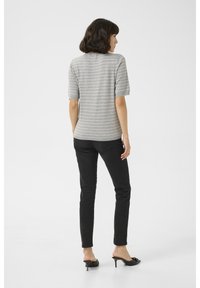 Vrouw met kort donker haar, gekleed in een grijs gestreept korte mouwen top, zwarte skinny jeans en zwarte muiltjes met hak, gekeerd met haar rug naar voren tegen een effen achtergrond.