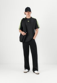 Sort oversized t-shirt med neongrønne striber, sorte vide bukser og hvide sneakers, accessorized med en sort taske og kasket.