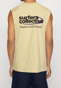 Person wearing a sleeveless beige shirt with "surfers collective" logo and "Brought to you by Originals" text on the back, paired with navy shorts.