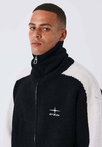 Homme aux cheveux courts et bouclés portant une veste zippée noire et blanche avec un col haut et un logo "PXP" sur la poitrine, tourné légèrement vers la gauche.