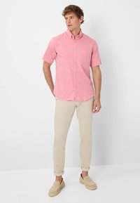 Camicia a maniche corte con bottoni in tessuto rosa chiaro con finitura testurizzata, dotata di una tasca sul petto. Abbinata a pantaloni beige slim-fit e scarpe slip-on.
