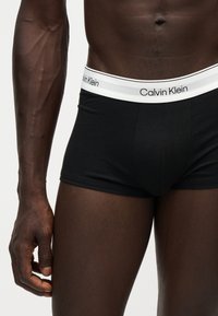 Άνδρας κορμός και άνω πόδια με μαύρα boxer Calvin Klein με λευκή ζώνη και ορατό όνομα μάρκας, σε ανοιχτό φόντο.