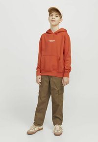 Felpa arancione con tasca frontale e logo bianco, abbinata a pantaloni cargo kaki con tasche laterali e sneakers beige con accenti crema.
