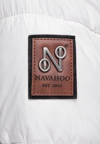 Bijela puffer jakna sa smešnim kožnim etiketom s urezanim metalnim logom "N" i "NAVAHOO EST 2014", obrubljena crnim materijalom.