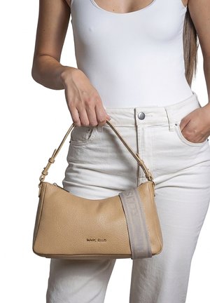 Donna con abito bianco che tiene una borsa in pelle texturizzata color cuoio Marc Ellis con tracolla regolabile e logo sul davanti.