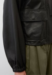 Giacca di pelle nera con colletto alto, tasca a toppa e dettagli dei polsini arricciati, abbinata a pantaloni plissettati verde oliva.