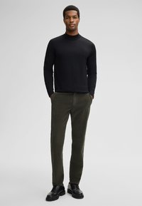 Zwarte coltrui, olijfgroene corduroy broek en zwarte veterschoenen. Model staat met handen in de zakken tegen een lichte achtergrond.
